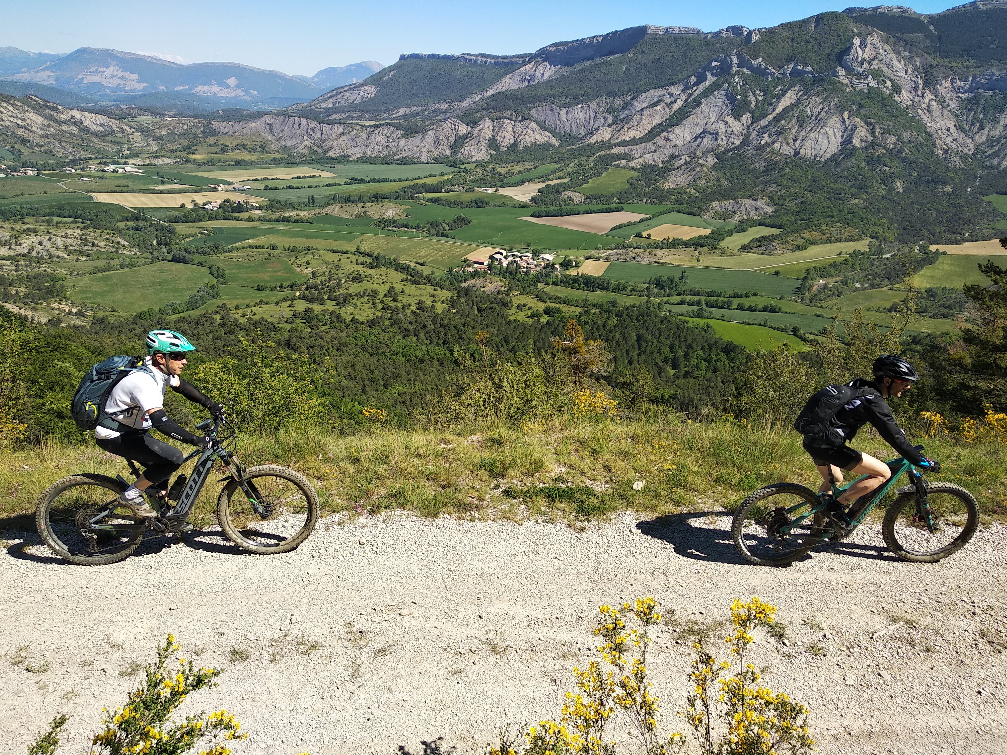 Two bikers cycle on electronic mountain bikes on a gravel path with bocage, pastured terrain with trees, in the background. This is in the French Alps on a sunny day on a sustainable cycling tour.