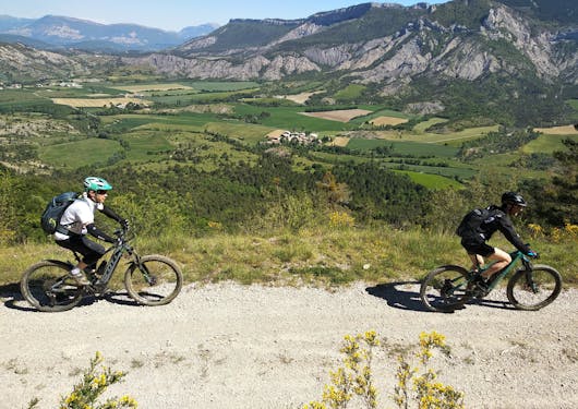 Two bikers cycle on electronic mountain bikes on a gravel path with bocage, pastured terrain with trees, in the background. This is in the French Alps on a sunny day on a sustainable cycling tour.