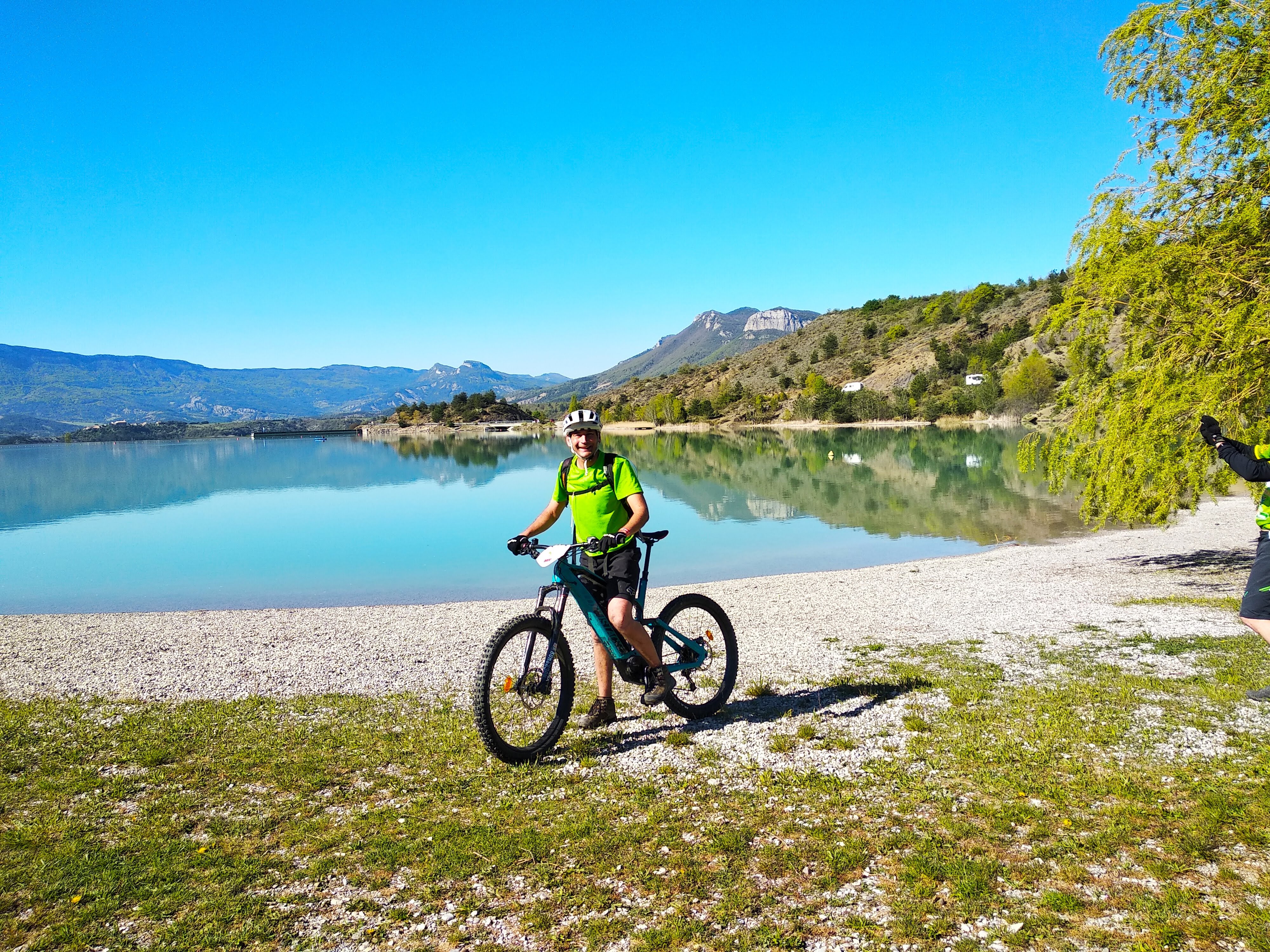 A cycler pauses, smiling at the camera straddling his bike, standing in front of a blue alpine lake. This is in the French Alps on a sunny day on a sustainable cycling tour.