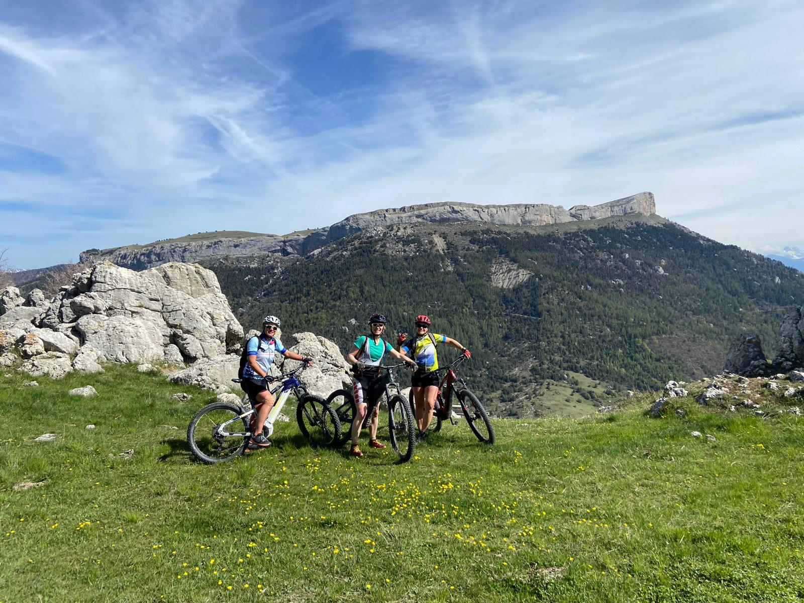 A group of 3 female bikers pause with mountains behind them as they rest on a grassy plateau. This is in the French Alps on a sunny day on a sustainable cycling tour.