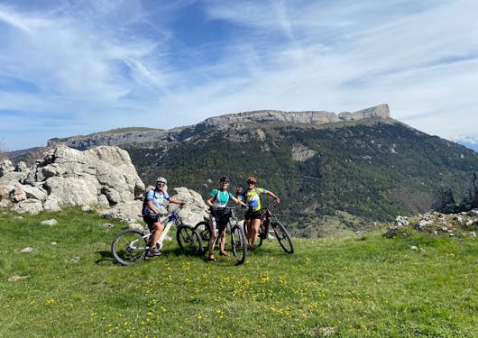 A group of 3 female bikers pause with mountains behind them as they rest on a grassy plateau. This is in the French Alps on a sunny day on a sustainable cycling tour.