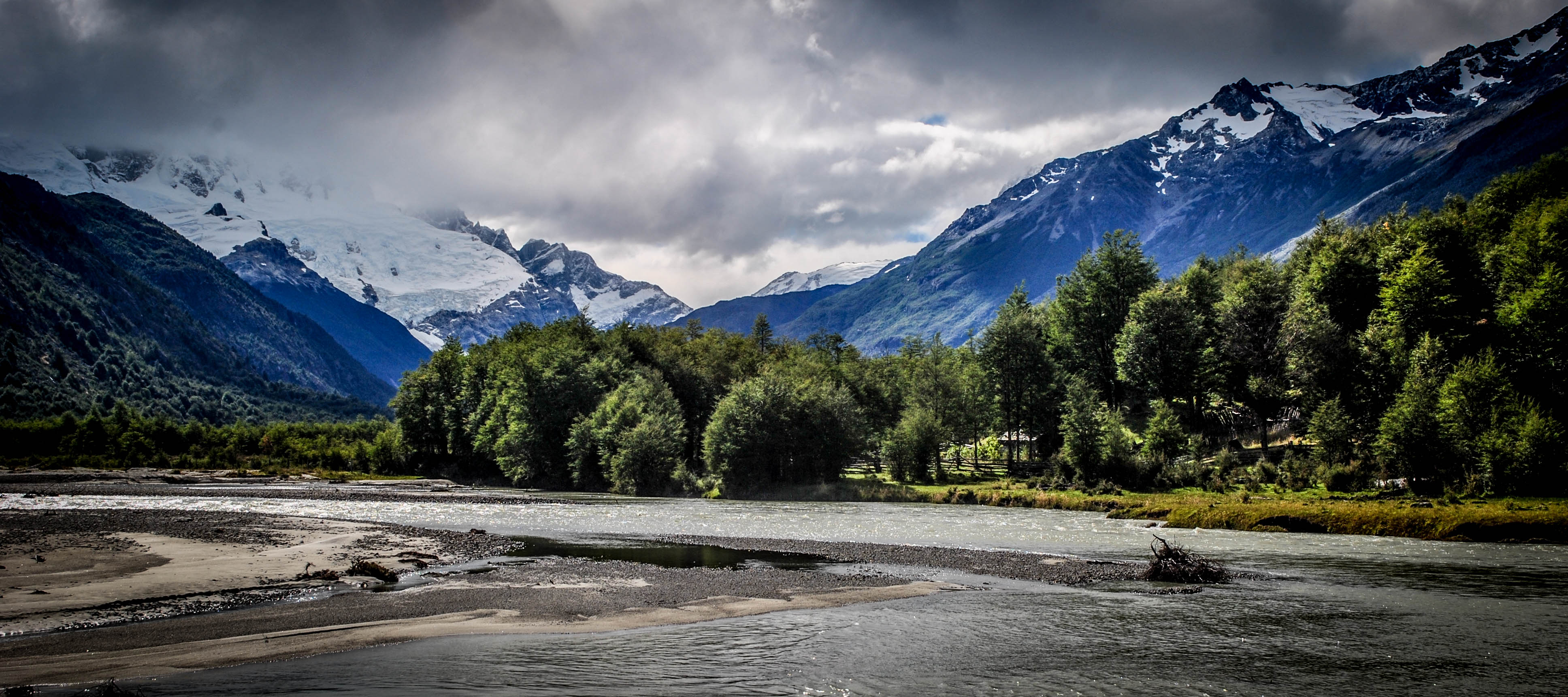 Flat terrain is in front of trees, with mountains covered by some clouds in the background. This is in the wild wilderness of Chilean Patagonia during a responsible supported trek along the El Gaucho Way.
