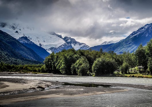 Flat terrain is in front of trees, with mountains covered by some clouds in the background. This is in the wild wilderness of Chilean Patagonia during a responsible supported trek along the El Gaucho Way.