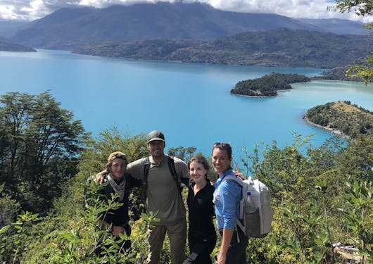 Two men and two women stand with their arms around each other, facing the camera and smiling along the Gaucho Way in Chilean Patagonia, with a bright blue lake behind them on a blue sky sunny day.
