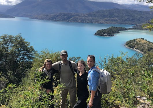 Two men and two women stand with their arms around each other, facing the camera and smiling along the Gaucho Way in Chilean Patagonia, with a bright blue lake behind them on a blue sky sunny day.