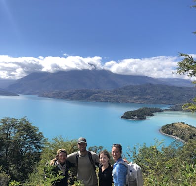 Two men and two women stand with their arms around each other, facing the camera and smiling along the Gaucho Way in Chilean Patagonia, with a bright blue lake behind them on a blue sky sunny day.