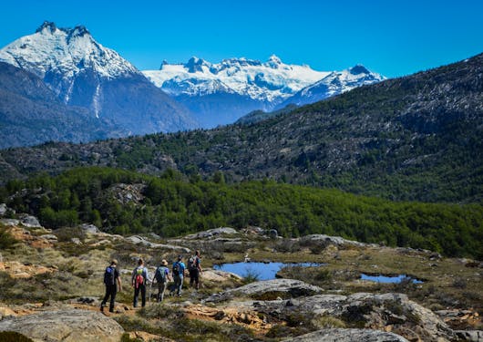 A group of 5 trekkers walk along the Gaucho Way in wild Chilean Patagonia on a responsible trekking tour. They walk over mixed terrain of dirt and rocks, with a small pond nearby and snow-capped mountains in the distance.