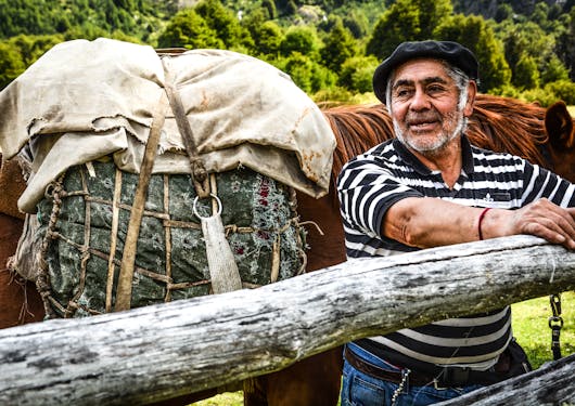 A local Chilean man with salt and pepper facial hair, a black and white striped t shirt, and a black cap stands in front of a brown horse behind a wooden fence railing in Patagonia along the Gaucho Way.