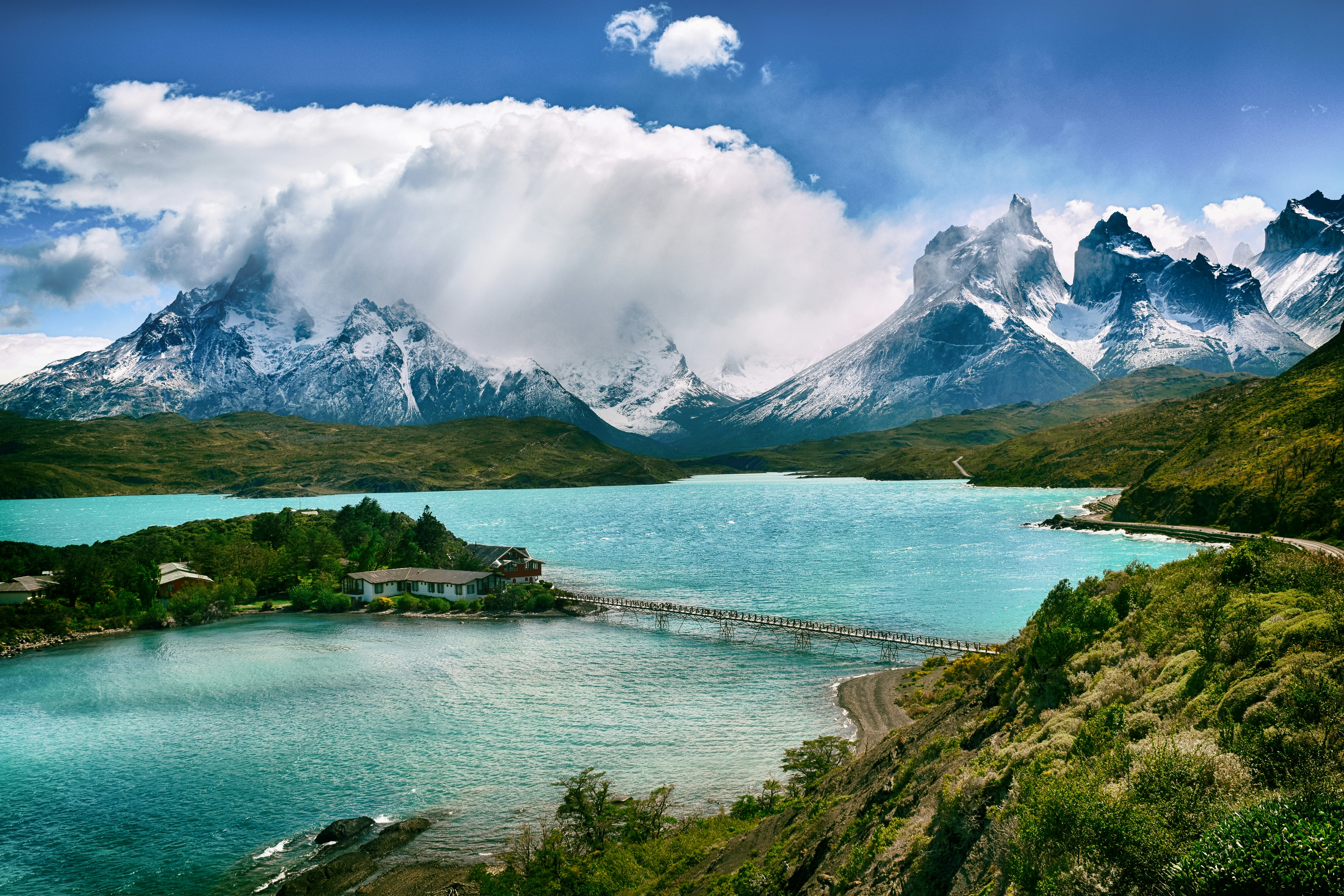 Torres del Paine National Park in Chile is shown from an aerial view with a teal blue lake surrounding a tree-filled island and a bridge connecting the two. Snow-covered mountains are in the background, with a bright blue sky and bright white clouds covering some of the peaks.