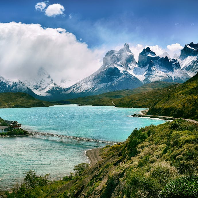 Torres del Paine National Park in Chile is shown from an aerial view with a teal blue lake surrounding a tree-filled island and a bridge connecting the two. Snow-covered mountains are in the background, with a bright blue sky and bright white clouds covering some of the peaks.