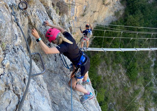 A climber faces a rock wall and she climbs rungs to the side, and another climber stands on a suspension bridge down below along a Via Ferrata route in the French Alps.