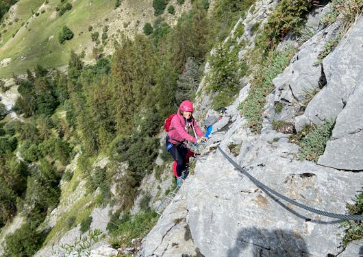 A climber comes up a metal rungs and holding a support rail as they climb a rock face along a Via Ferrata route in the French Alps with trees and green land below.