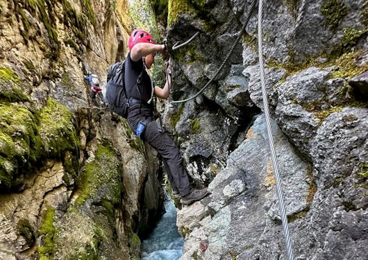 A climber stands on a rock face and holds metal rungs for support as a stream rushes below along a Via Ferrata route in the French Alps.