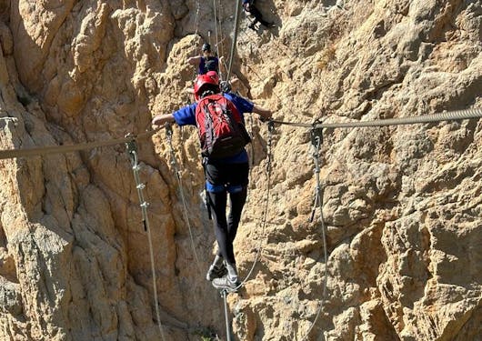A climber walks a metal tight rope bridge while holding support rails along a Via Ferrata route in the French Alps.