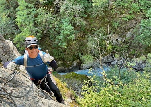 A man in hat and sunglasses leans against a rock face with greenery and a stream in the background and below him, along a Via Ferrata route in the French Alps on a sunny day in France.