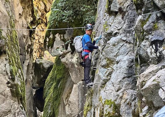 A climber stands on metal rungs, holding other metal rungs for support, on a rock face just above a rushing stream along a Via Ferrata route in the French Alps.