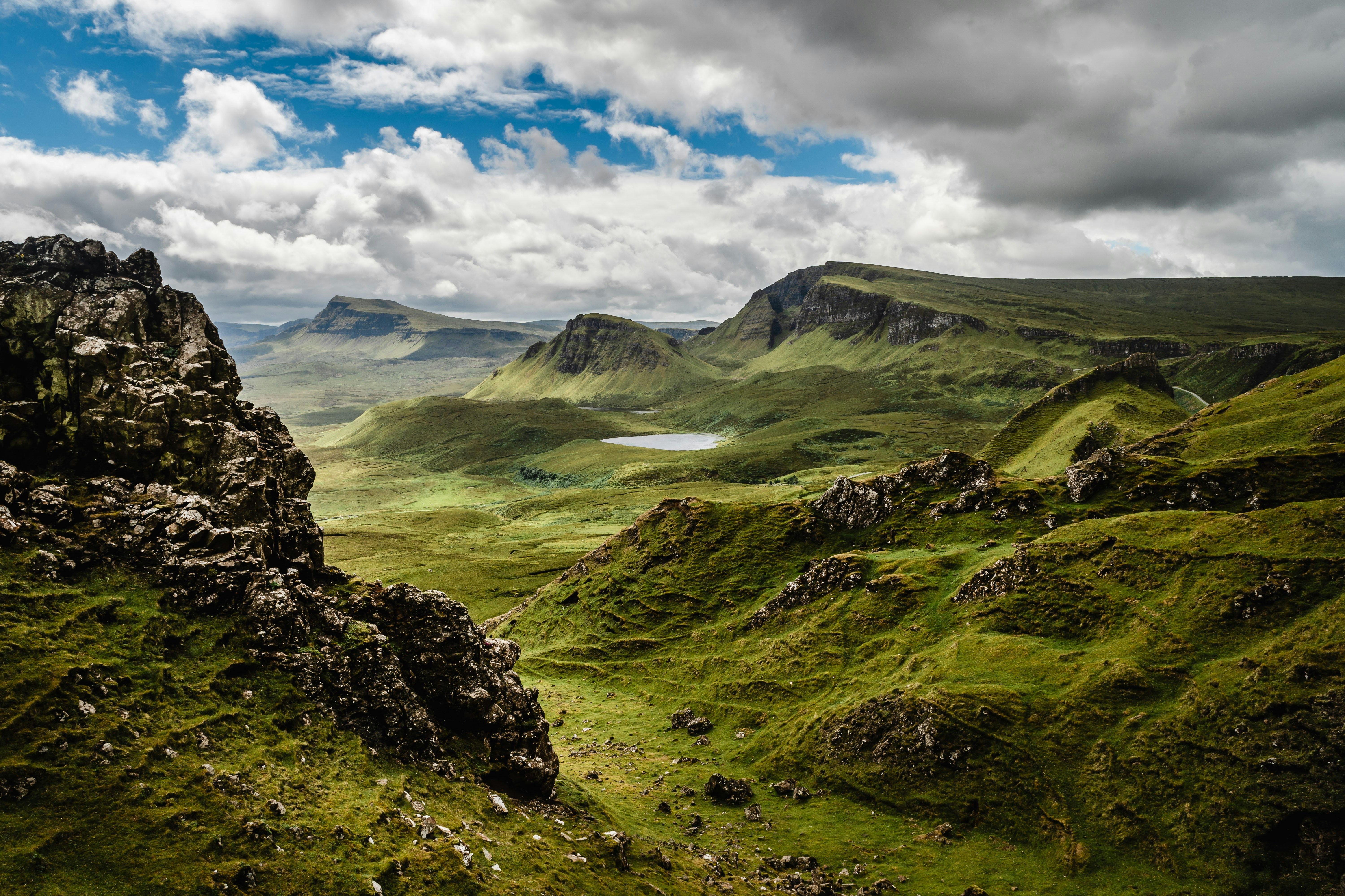 The Isle of Skye is shown in Scotland with mossy, craggy green hills and a cloudy sky with some blue peeking through.