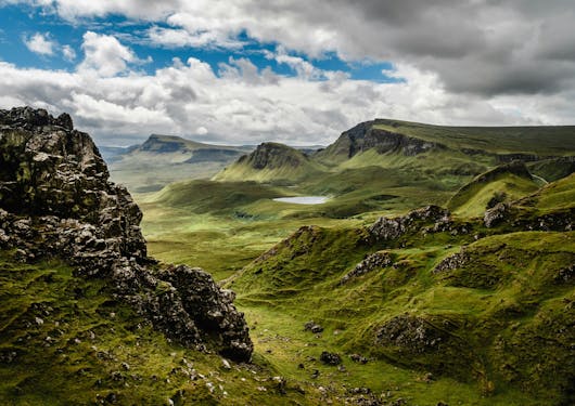 The Isle of Skye is shown in Scotland with mossy, craggy green hills and a cloudy sky with some blue peeking through.