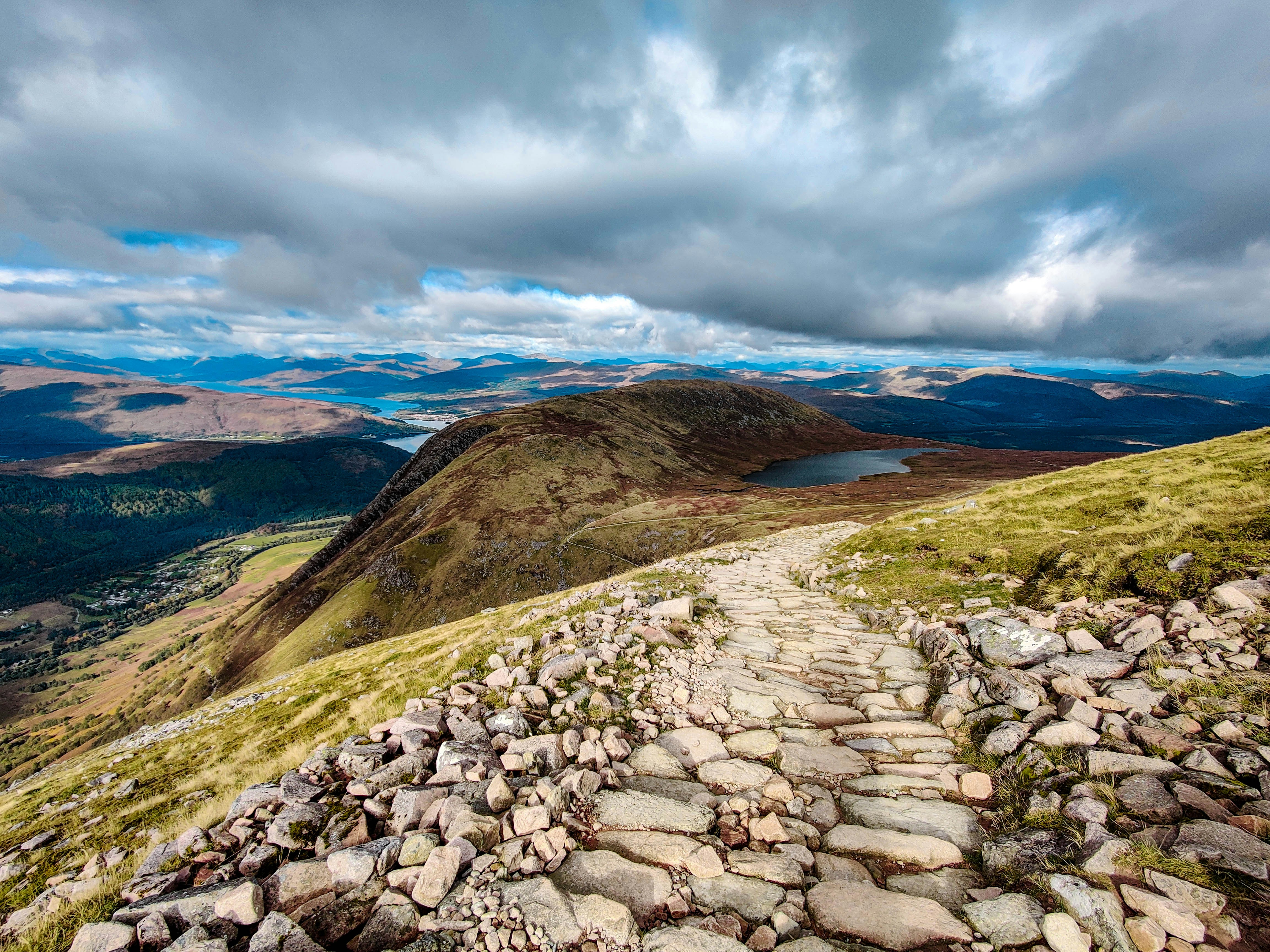 The view from Ben Nevis, Scotland's tallest mountain, near Fort William, the capital of the Scottish Highlands, is shown with a rocky path extending out and hills in the background.