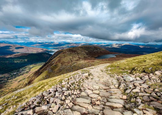 The view from Ben Nevis, Scotland's tallest mountain, near Fort William, the capital of the Scottish Highlands, is shown with a rocky path extending out and hills in the background.
