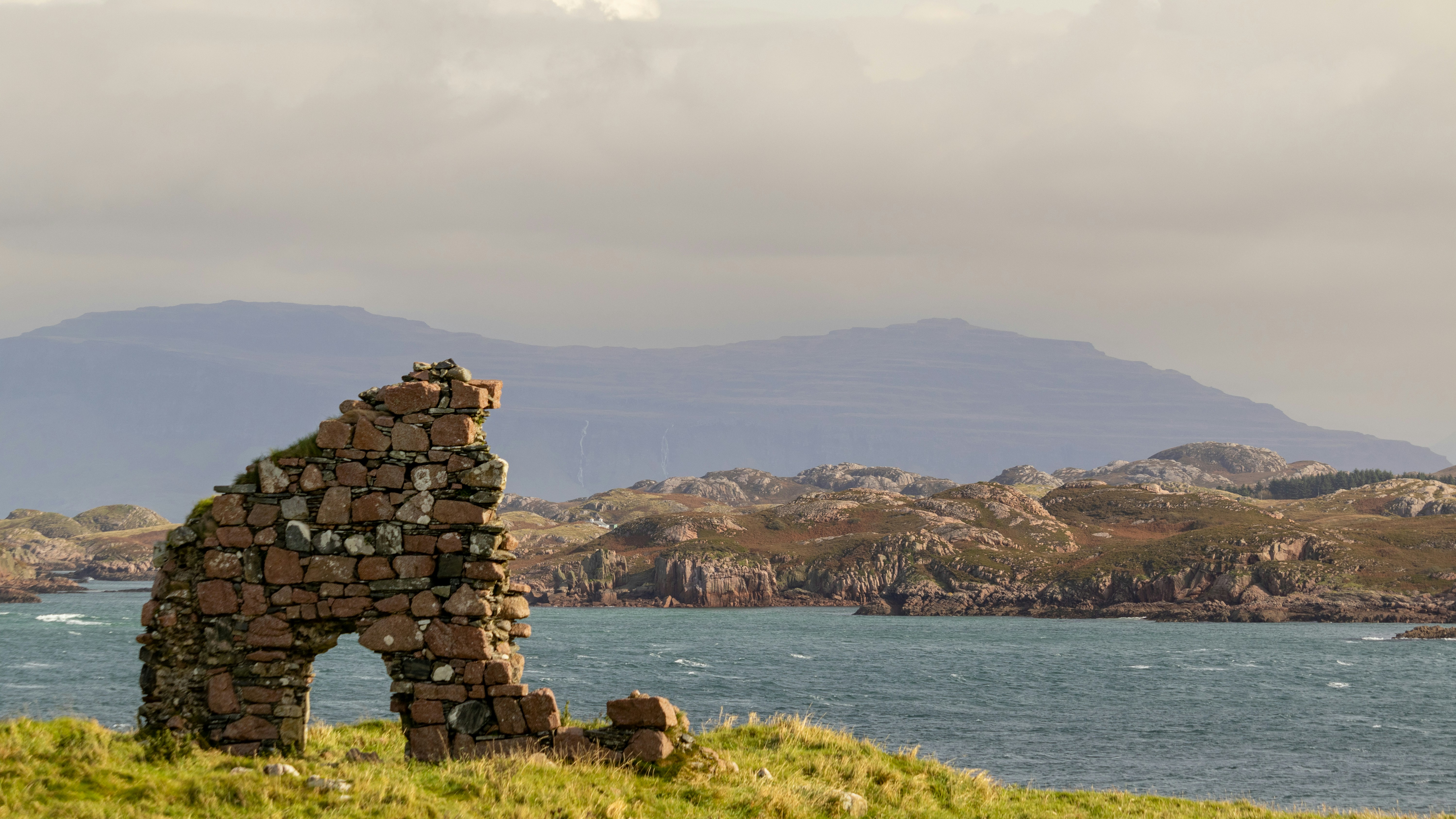 A view of the Isle of Iona off of Scotland's west coast is shown, with rock remnants forming an arch and hilly terrain in the background beyond the water.