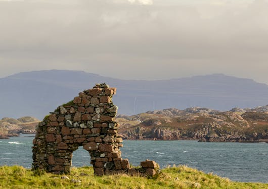 A view of the Isle of Iona off of Scotland's west coast is shown, with rock remnants forming an arch and hilly terrain in the background beyond the water.