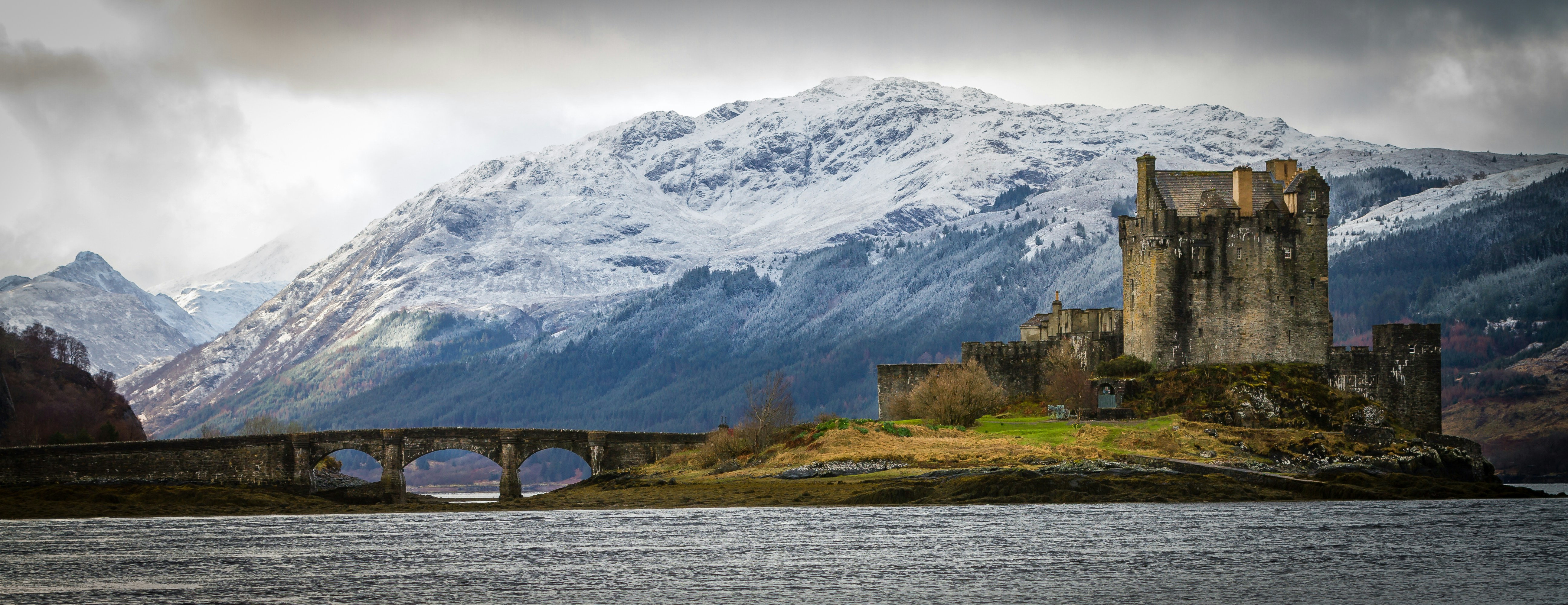 The famously iconic Eilean Donan Castle in Scotland is shown across the water with a snow-capped mountain in the background.