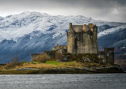 The famously iconic Eilean Donan Castle in Scotland is shown across the water with a snow-capped mountain in the background.