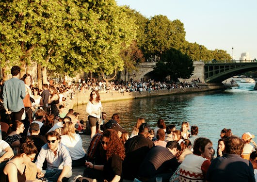 A crowd of people are sitting along the banks the Seine in Paris. You can see as the river bends crowds of people filling the entire river wall. In the distance you can see a bridge and you can also find trees lining the river. 