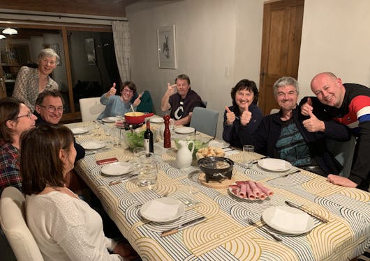 A group of 9 middle-aged to senior-aged adults sit around the dining room table at a guesthouse in the southern French Alps where they're staying on a sustainable self-guided walking holiday of the area.
