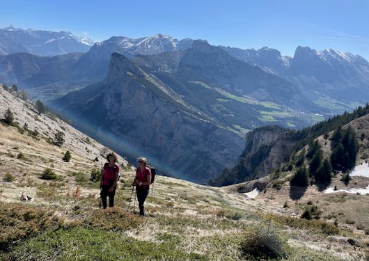 Two adult ladies hike in the Devoluy Massif area of the southern French Alps on a sunny day, with peaks in the background.