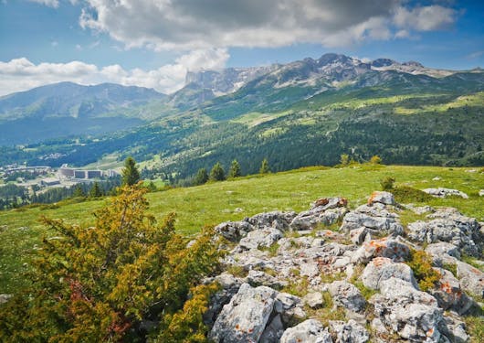 A view of limestone rocks, green grass, and mountain peaks from along a trail in the Devoluy Massif area of the southern French Alps on a sunny day with some clouds overhead.