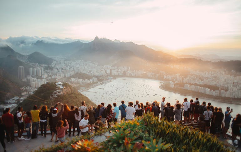 A line of people lean against a barrier admiring the view from a mountain top overlooking a city below with hills in the distance during sunset
