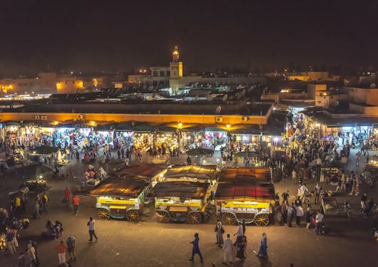 A night market, or souk, is shown in Marrakesh Morocco, from a distance. It's lights are twinkling from the stalls of many vendors.