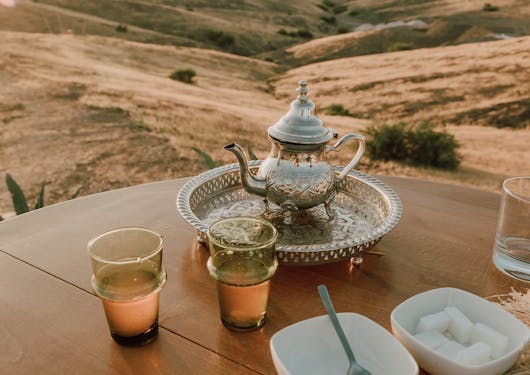 A table is set up in Morocco serving traditional tea in a kettle with 2 glasses nearby, with a bowl of sugar and another bowl with a spoon in it.
