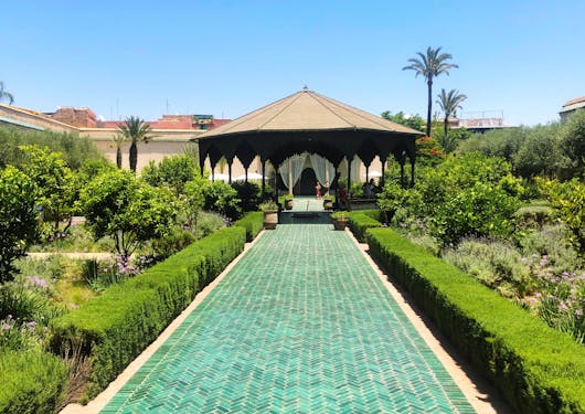 A secret garden is shown, looking down a long green bring path that ends in a gazebo. Greenery lines the brick path and it looks lush. It's a blue sky sunny day in Morocco.