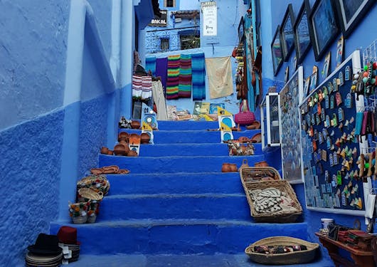 The famous Blue City of Morocco, also known as Chefchaouen, is shown from the vantage point of the bottom of some steps, looking up. The steps are lined with various colorful handicrafts and woven goods.