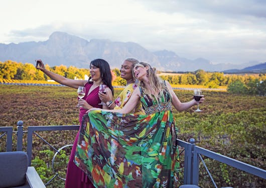 Three women with different skin tones wearing colorful dresses smile together while taking a selfie in the South African Cape Winelands, while holding glasses of wine.