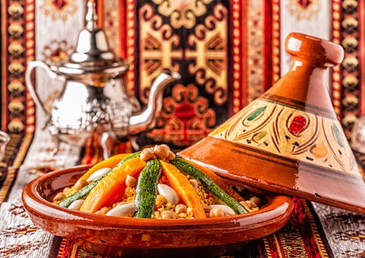 A colorful vegetarian Tagine dish is shown in Morocco with bright reds, yellows, and greens, with a silver tea pot in the back and placed against red and orange intricately woven fabric.