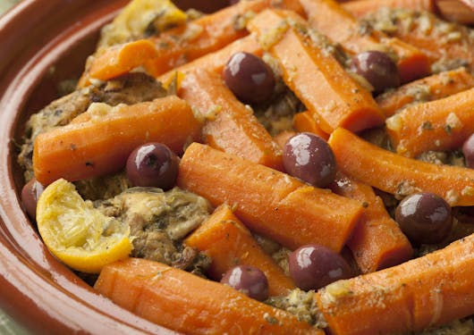 A Moroccan dish in traditional rust colored earthenware is shown; it contains chickens and orange carrots and dark olives with lemon wedges.
