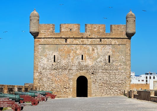 A fort in Essaouira, Morocco is shown. It has a blocky structure and is against a bright blue, cloudless sky.