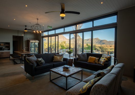 An inside view of the Cheetah Project's Volunteer House accommodations in South Africa. Couches surround a table and a big window with greenery and small pointed hills are in the background.