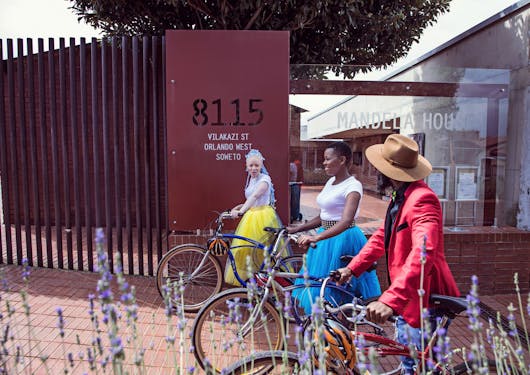 Visiting Nelson Mandela's house on Vilakaze Street in Soweto, Johannesburg, South Africa. Three adult women are on bikes in front of the address, one is wearing a yellow skirt and white top, one a blue skirt and white top, and a foremost person is wearing a red long sleeve jacket and a hat.