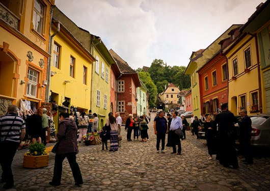 The colorful town of Sighisoara in Romania boasts houses in yellows, greens, reds, and oranges. People mill about the street beneath an overcast sky.