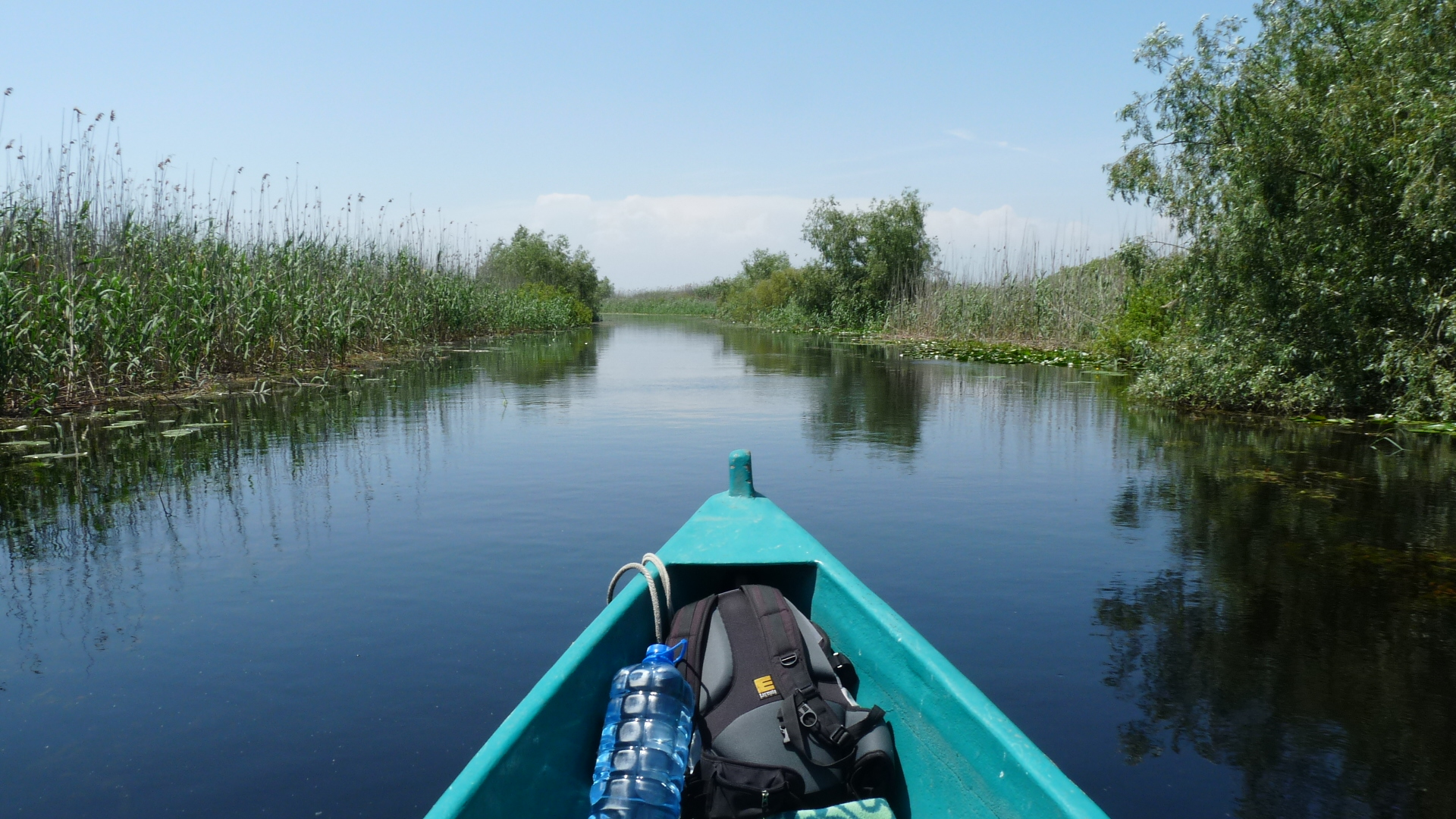 A teal boat heads down Romania's Danube Delta River with a backpack and a giant plastic water jug in the front, and greenery on both sides as well as in front of it. It's a blue sky day.