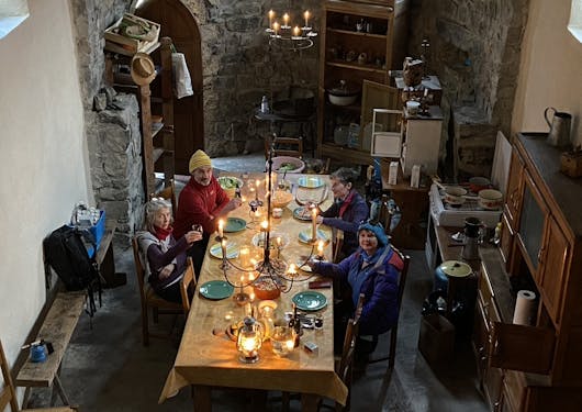 A group of 4 adults sit around a dinner table with candles lit, as seen from above while they enjoy a meal together at the old church refuge accommodations in the French Alps.