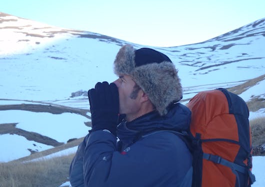 A man in a blue parka, fur hat, and red backpack howls to the wolves on a snowy hillside in the French Alps.