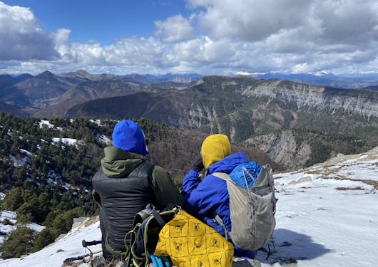 Two adult onlookers bundled up for cold weather in blue and black parks, and yellow and blue hats, sit on a snowy outlook gazing out over the landscape of the French Alps.