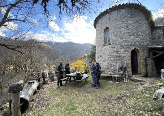 A church refuge provides accommodation in the French Alps to a select group of visitors learning about responsible wolf tracking from expert guides. A group of 6 or 7 adults stand out front around a wooden picnic table in the daylight.