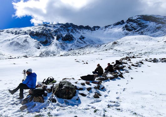 A group of adults sit atop boulders on a snowy plateau as they rest mid-day on the responsible wolf tracking tour in the French Alps.